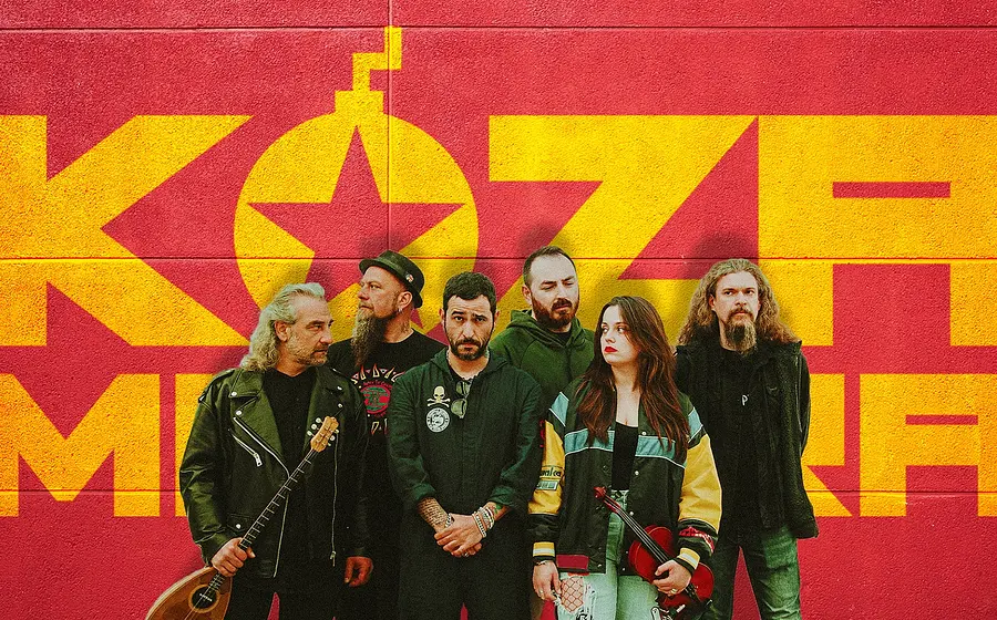 Sechs Personen stehen vor einer rot-gelben Wand mit großem ‚KOZA MOSTRA‘‑Schriftzug; einige halten Musikinstrumente wie Bouzouki und Geige, alle tragen legere bis rockige Kleidung