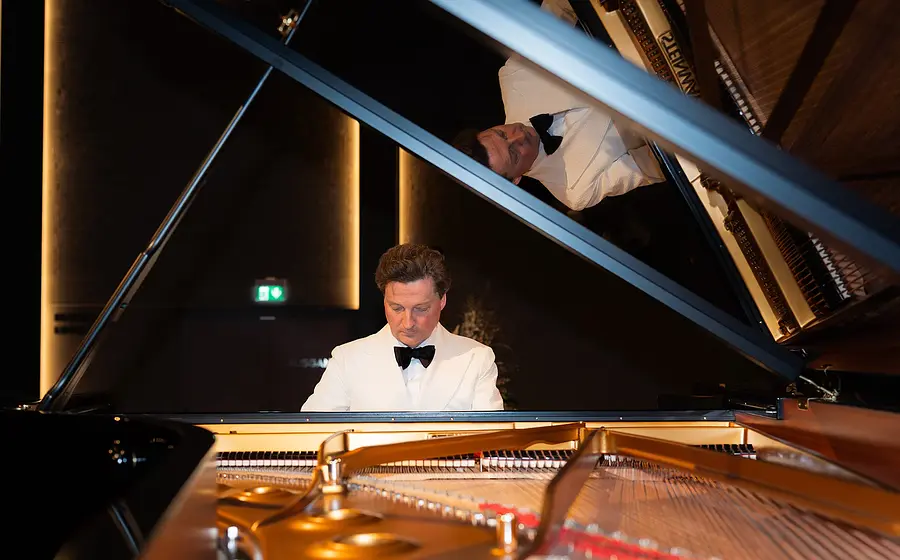 Ein Pianist im weißen Smoking mit schwarzer Fliege an einem geöffneten Konzertflügel, aufgenommen durch den Flügelrahmen mit Spiegelung im aufgeklappten Deckel, vor warm beleuchtetem Konzertsaalhintergrund.