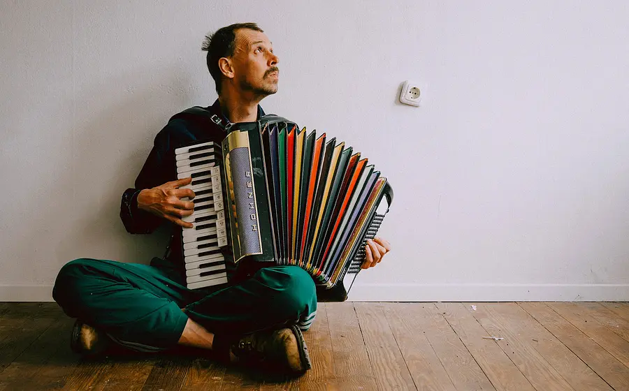 Foto von Maxi Pongratz in grüner Hose zur Seite Seite blickend mit einem bunten Akkordeon auf einem hellen Holzboden sitzend, vor hellem Hintergrund
