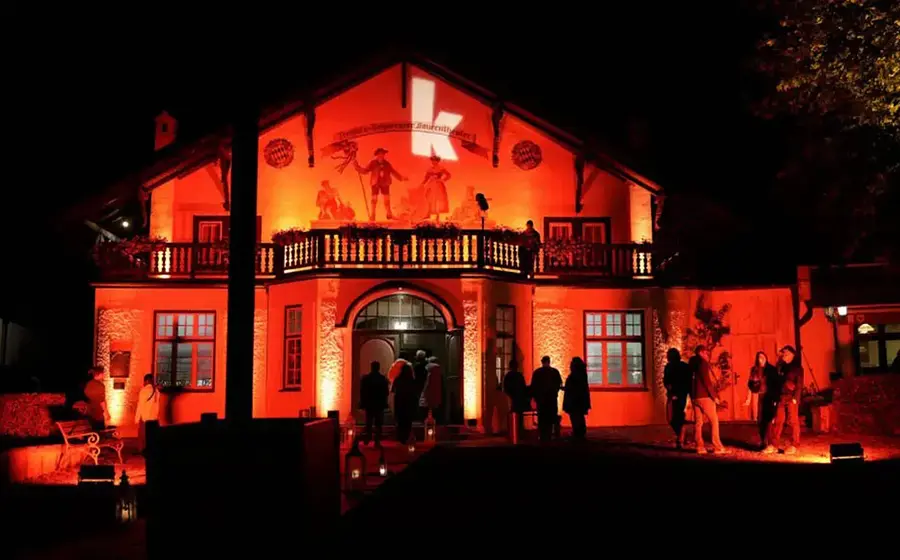 Beleuchtetes Gebäude bei Nacht in rotem Licht mit Balkon und Eingang, davor mehrere Personen; an der Fassade ein großes beleuchtetes K.