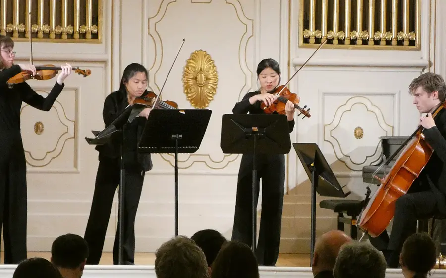 Vier Musiker in schwarzer Konzertkleidung spielen auf einer hellen Bühne mit weiß-goldenem Saalinterieur. Violinen, Viola und Cello sowie die warme Beleuchtung verleihen dem Konzert eine elegante, klassische Atmosphäre.