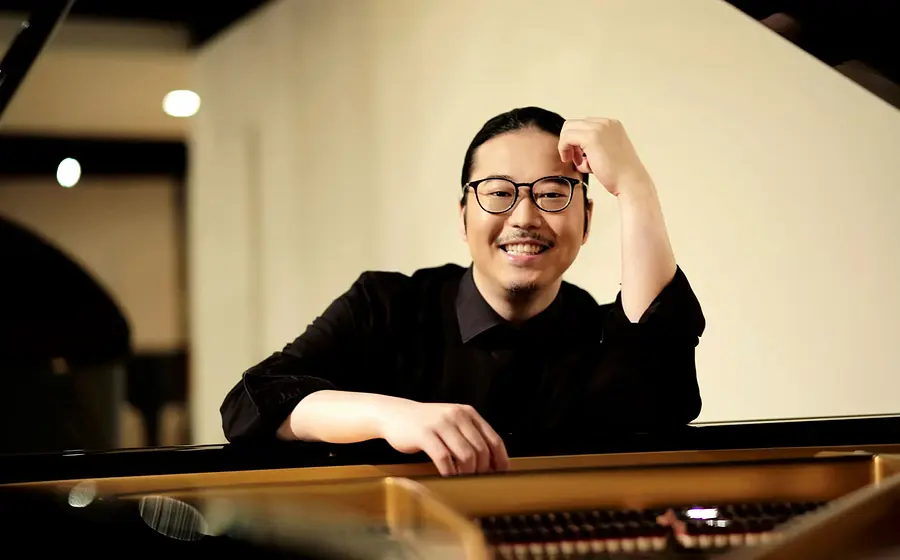Porträt des Pianisten Kyohei Sorita an einem Flügel, halbnah aufgenommen, lehnt sich mit einem Arm auf das Instrument, trägt dunkle Kleidung und sitzt in einem Innenraum mit weicher Beleuchtung im Hintergrund.
