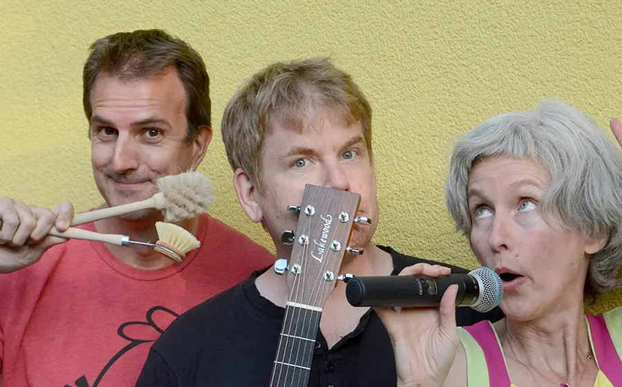 Pressefoto von La Psychotta mit drei Mitwirkenden vor gelber Wand, eine Person hält Mikrofon und hebt die Hand, eine weitere hält eine Gitarre vor das Gesicht und die dritte verschiedene Bürsten und Alltagsgegenstände in die Kamera.