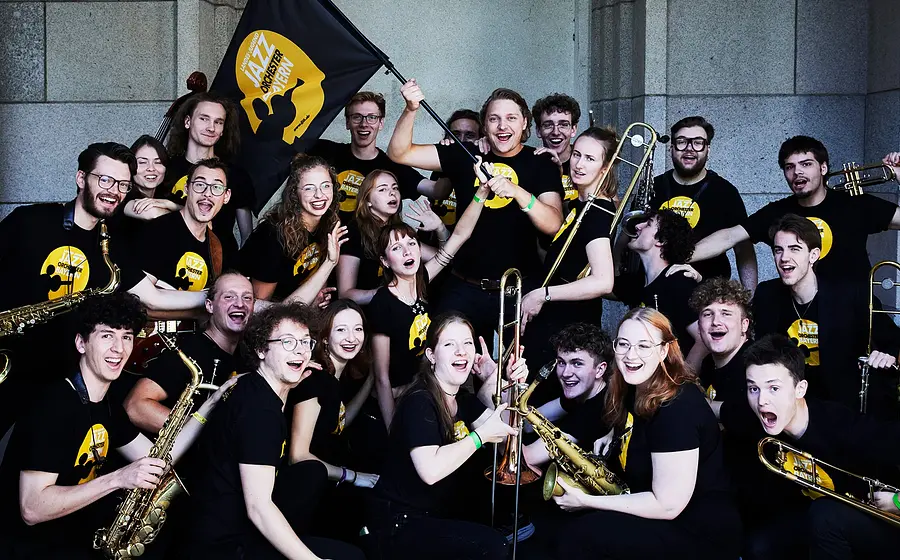 Eine große Gruppe junger Musikerinnen und Musiker posiert mit Saxofonen, Trompeten und Posaunen. In der Mitte wird eine Fahne des Landesjugendjazzorchesters Bayern hochgehalten. Die Stimmung ist fröhlich und ausgelassen.