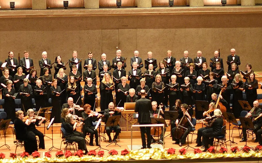 Großer Konzertchor in schwarzer Kleidung singt gemeinsam auf einer Bühne. Im Vordergrund dirigiert eine Person ein Kammerorchester mit Streichinstrumenten, während der Chor in mehreren Reihen hinter dem Ensemble aufgestellt ist.
