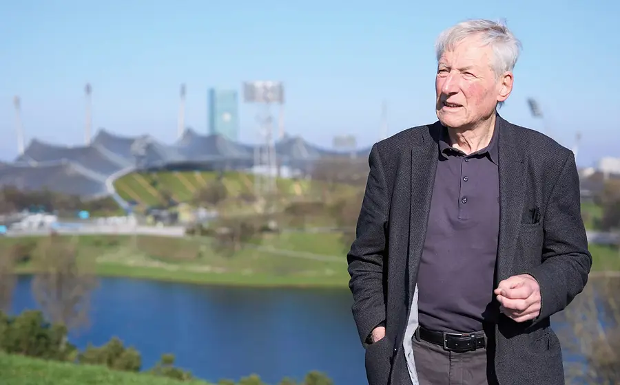 Person steht im Freien vor einer StadtlandArchitekt Prof. Fritz Auer steht im Freien vor dem Olympiagelände in München mit See und Stadion im Hintergrund bei sonnigem Wetterschaft mit See und Olympiastadion im Hintergrund, bei sonnigem Wetter