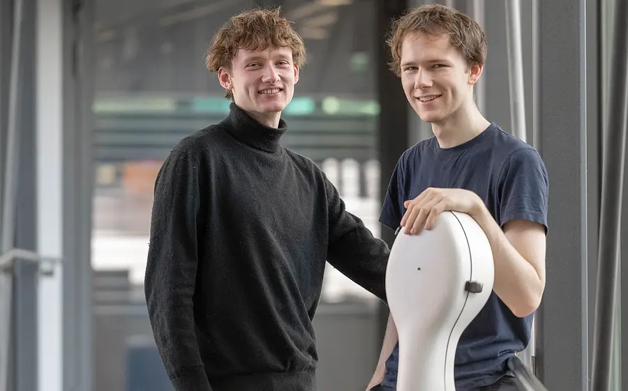 Zwei Musiker stehen in einem modernen Gebäude. Eine Person lehnt an einem Violoncello-Koffer, die andere steht daneben