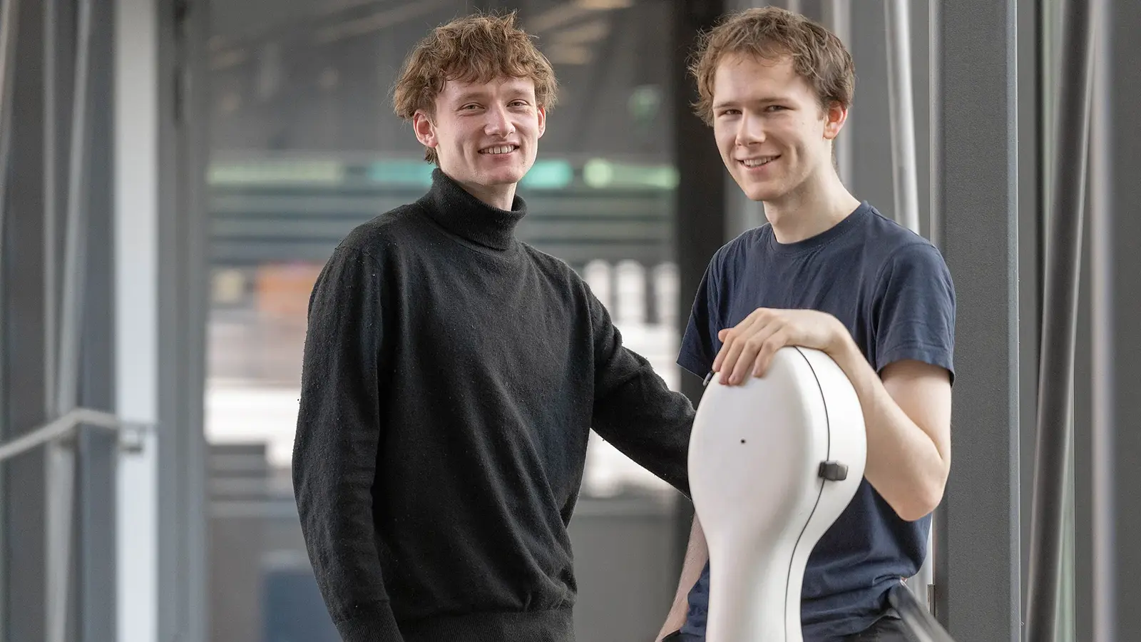 Zwei Musiker stehen in einem modernen Gebäude. Eine Person lehnt an einem Violoncello-Koffer, die andere steht daneben