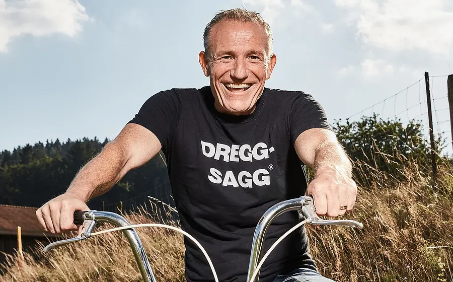 Porträt von Michl Müller: lachend auf einem Fahrrad im Freien, mit schwarzem T-Shirt vor ländlicher Landschaft.