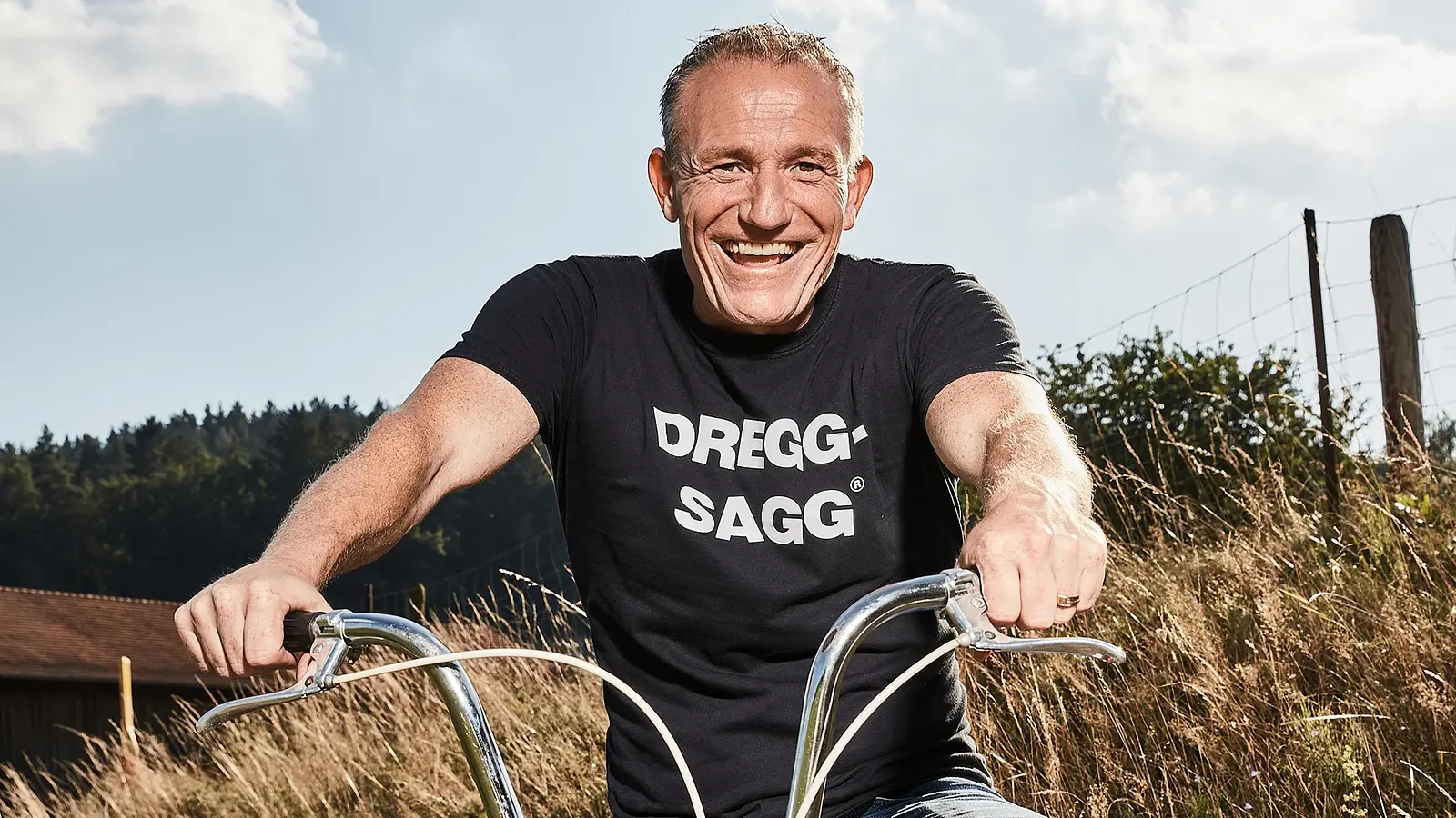 Porträt von Michl Müller: lachend auf einem Fahrrad im Freien, mit schwarzem T-Shirt vor ländlicher Landschaft.