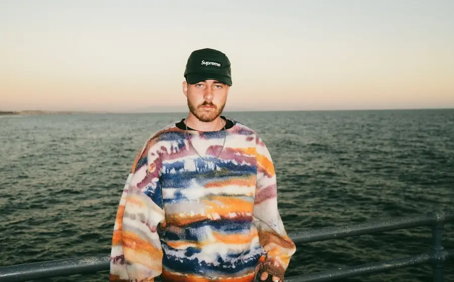 Eine Person in farbigem gemusterten Pulli und schwarzer Kappe steht an einer Brüstung, im Hintergrund das Meer
