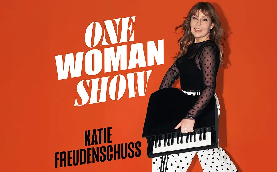 Poster der Kabarettistin Katie Freudenschuss vor orangefarbenem Hintergrund sie hält ein tragbaren Flüge mit den Aufschriften One Woman Show und Katie Freudenschuss links neben ihr