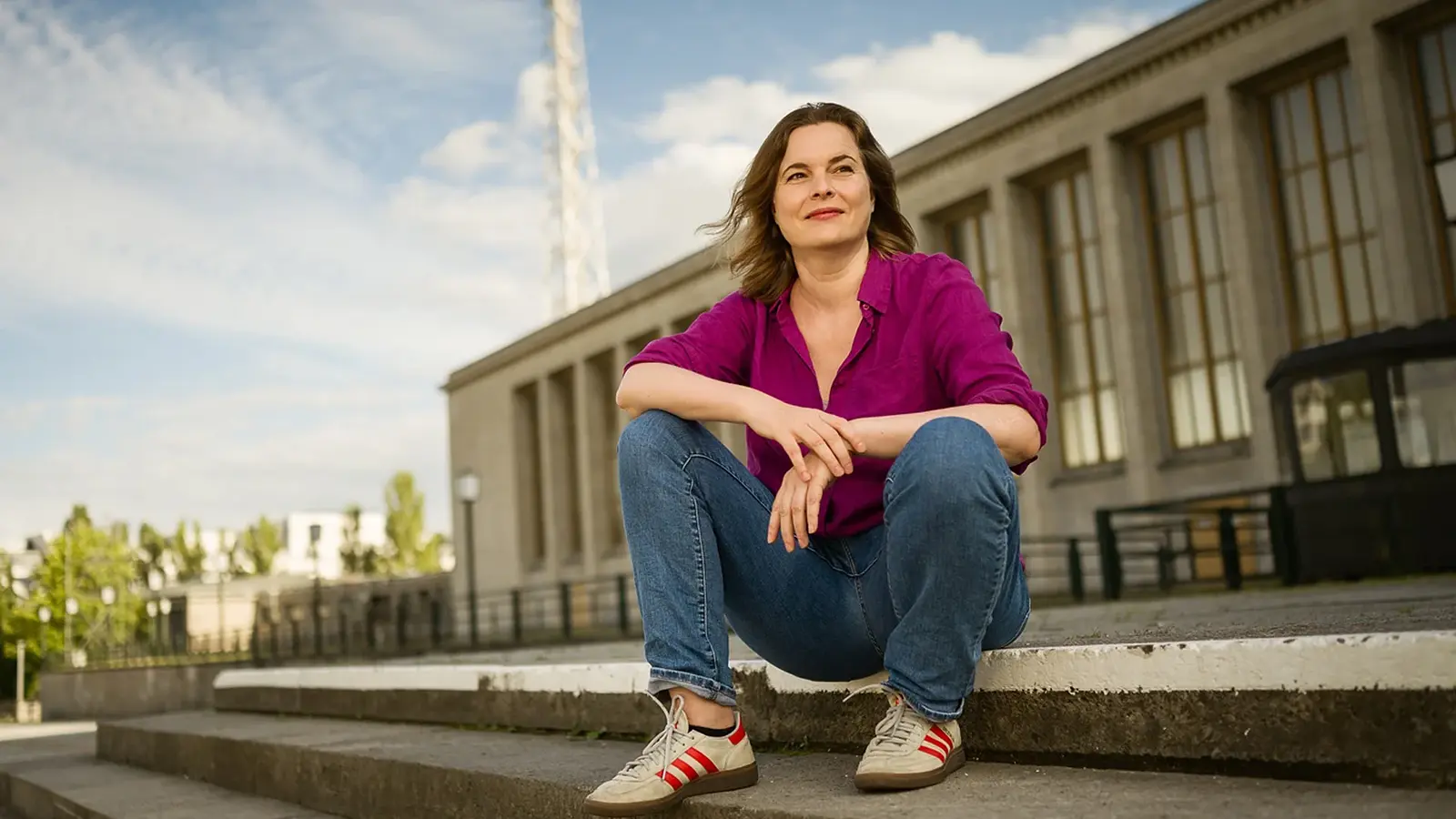 Katja Berlin sitzt draußen auf einer Treppe vor einem Gebäude und lächelt.