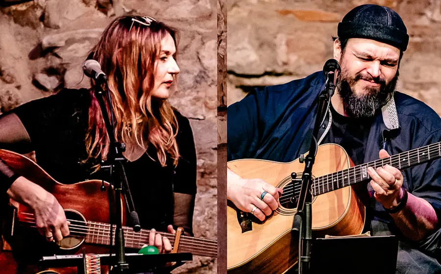 Zwei Personen mit Akustikgitarren bei einem Bühnenauftritt vor einer rustikalen Steinwand. Warme Braun- und Beigetöne sowie schwarze Kleidung schaffen eine intime Konzertatmosphäre.