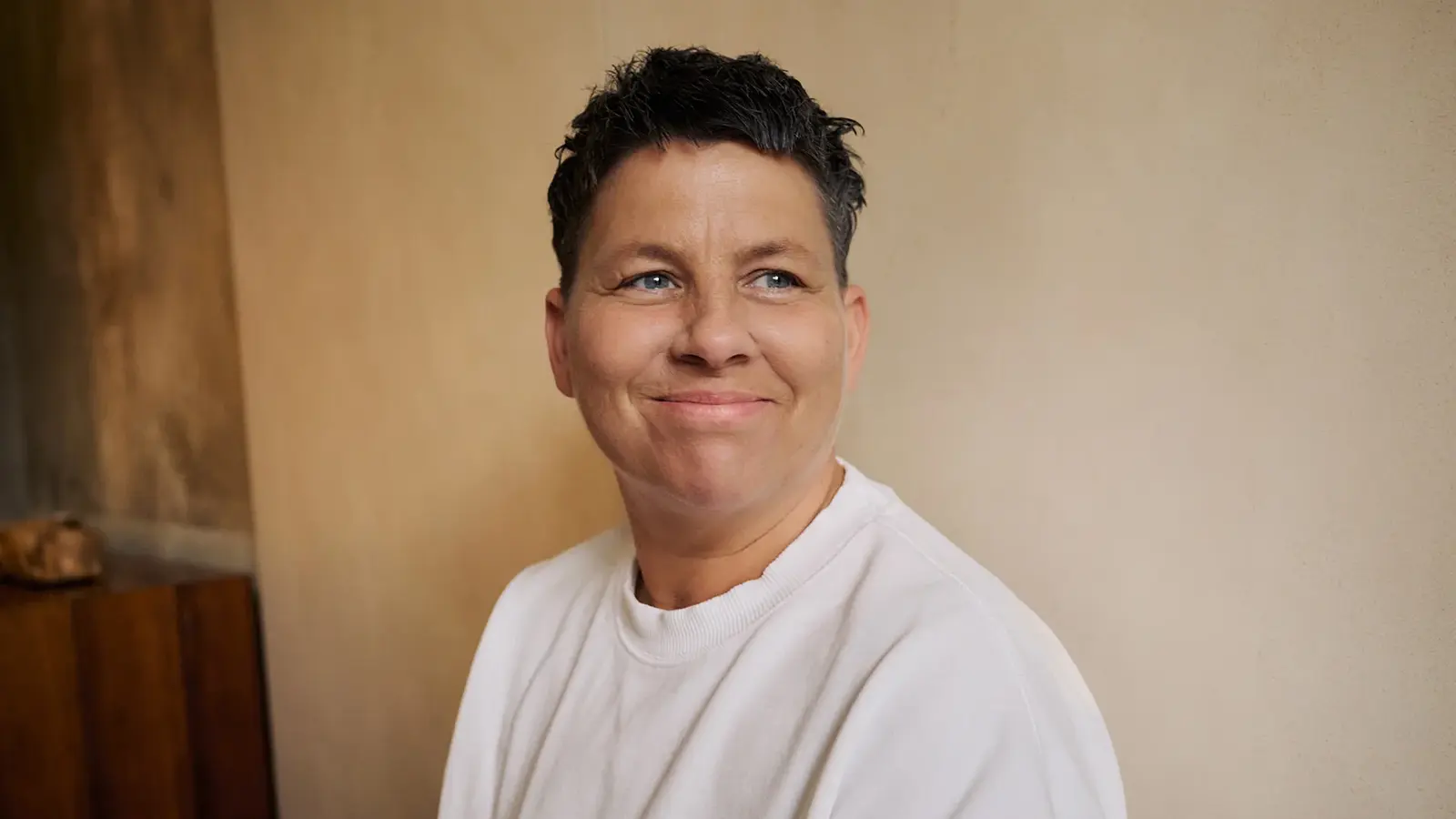 Kerstin Ott in weißem T-Shirt lächelnd, vor einer hellen Wand, auf der linken Seite eine Holzvertäfelung