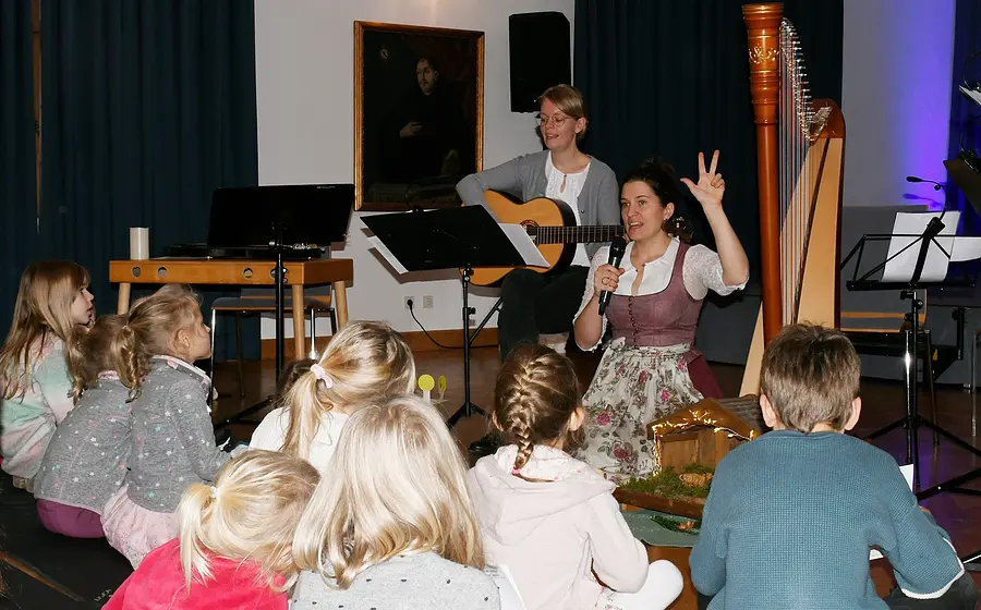 Kinder sitzen vor zwei Musikerinnen. Eine Frau im Dirndl spricht mit Mikrofon und erhobener Hand, die andere begleitet mit Gitarre. Daneben stehen eine Konzertharfe, Notenständer und eine kleine Krippe