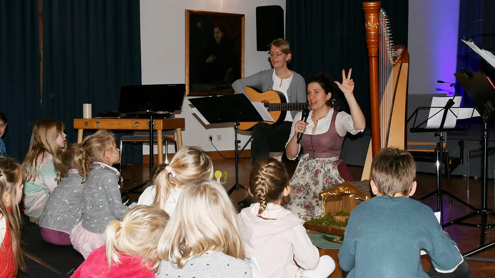 Kinder sitzen vor zwei Musikerinnen. Eine Frau im Dirndl spricht mit Mikrofon und erhobener Hand, die andere begleitet mit Gitarre. Daneben stehen eine Konzertharfe, Notenständer und eine kleine Krippe
