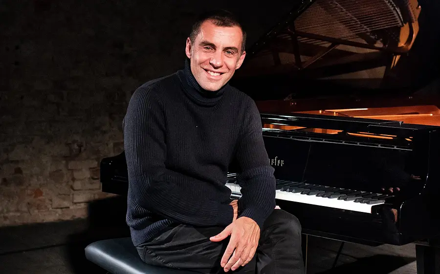 Pianist sitzt lächelnd an einem Flügel auf einer Bühne, Porträtfoto in dunkler Konzertatmosphäre.