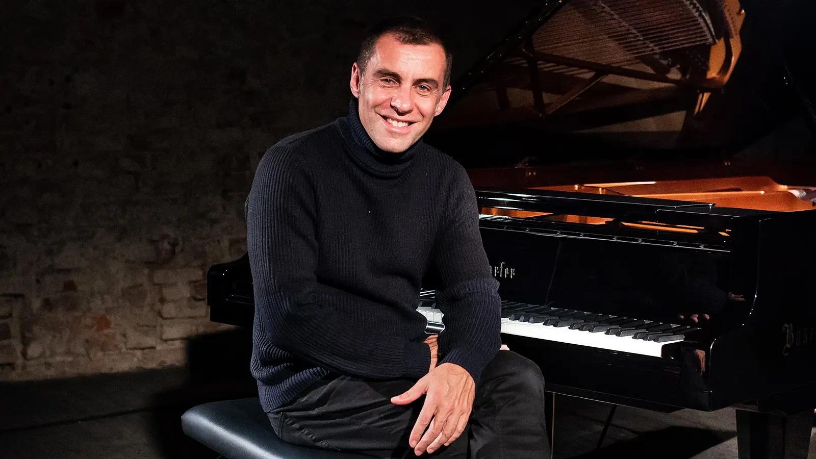 Pianist sitzt lächelnd an einem Flügel auf einer Bühne, Porträtfoto in dunkler Konzertatmosphäre.