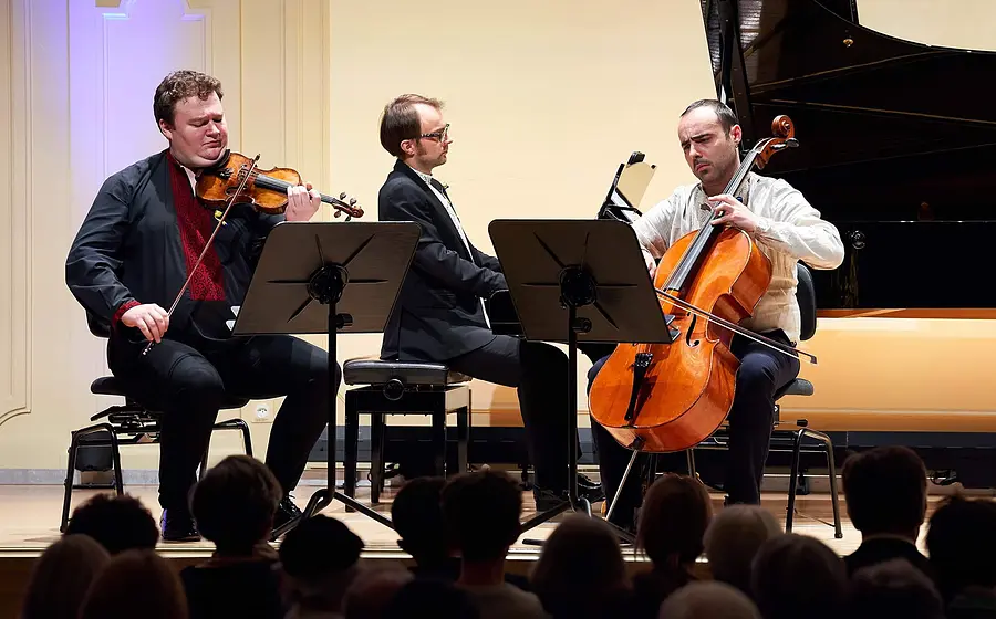 Drei Musiker bei einem Kammerkonzert auf einer Bühne: Violine, Klavier und Violoncello. Im Vordergrund ist das Publikum zu sehen, im Hintergrund ein Konzertflügel.