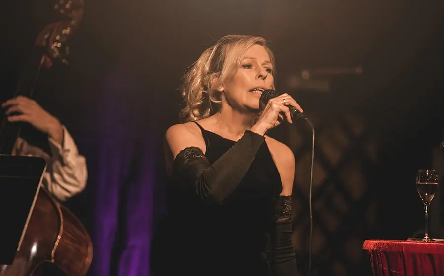 Bühnenfoto einer Sängerin im schwarzen Abendkleid mit Mikrofon auf einer Jazzbühne, begleitet von einem Kontrabass im Hintergrund, bei warmem Bühnenlicht neben einem kleinen Tisch mit Weinglas.