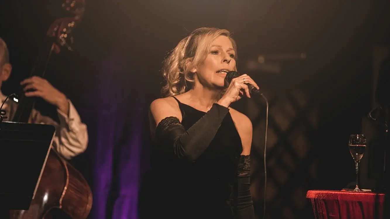 Bühnenfoto einer Sängerin im schwarzen Abendkleid mit Mikrofon auf einer Jazzbühne, begleitet von einem Kontrabass im Hintergrund, bei warmem Bühnenlicht neben einem kleinen Tisch mit Weinglas.