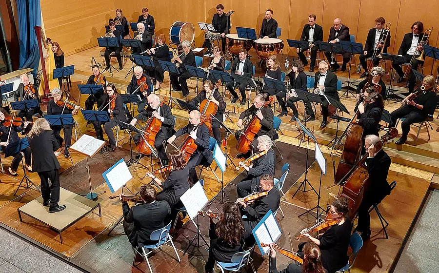 Bühnenfoto eines Symphonieorchesters mit Dirigentin oder Dirigent auf einem Podest vor großem Ensemble aus Streichern, Holz- und Blechbläsern sowie Pauken, Harfe und Schlagwerk, auf einer Konzertbühne mit hölzerner Wandverkleidung und zahlreichen Notenständern