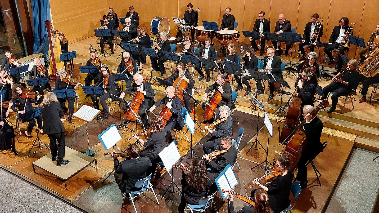 Bühnenfoto eines Symphonieorchesters mit Dirigentin oder Dirigent auf einem Podest vor großem Ensemble aus Streichern, Holz- und Blechbläsern sowie Pauken, Harfe und Schlagwerk, auf einer Konzertbühne mit hölzerner Wandverkleidung und zahlreichen Notenständern