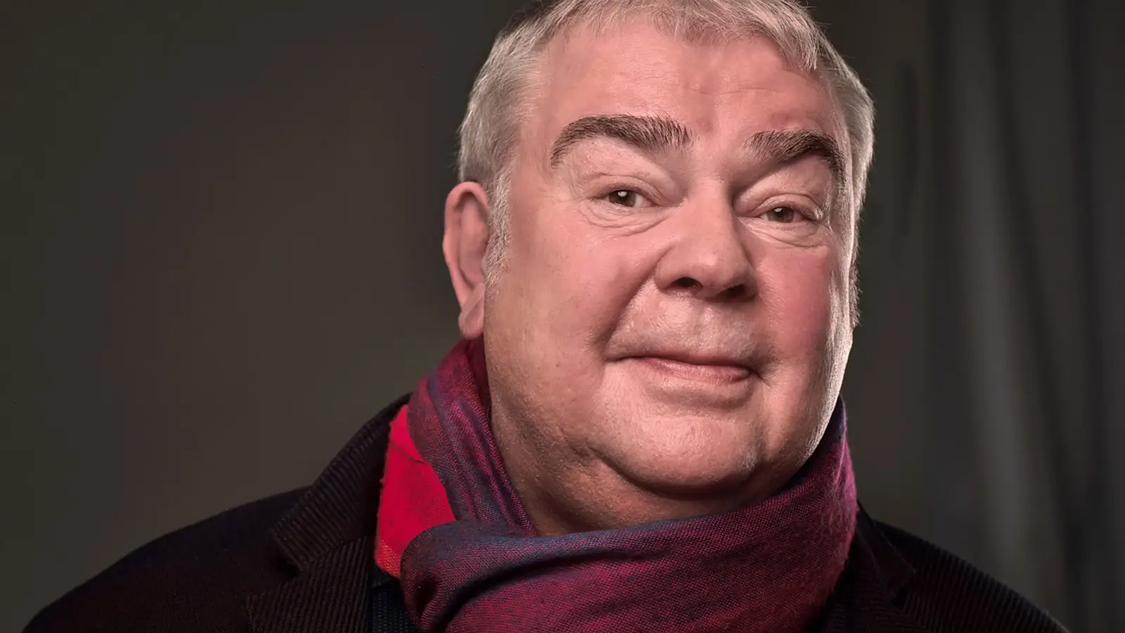 Portrait von Max Goldt mit rot-blauem Schal vor grauem Hintergrund