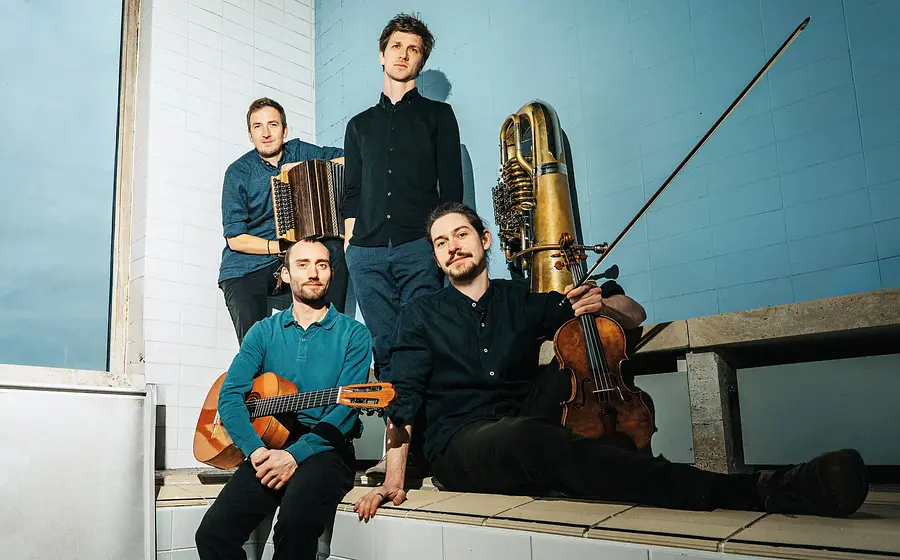 Vier Musiker mit Geige, Steirischer Harmonika, Gitarre und Tuba posieren in einem hellen Raum mit gefliesten Wänden