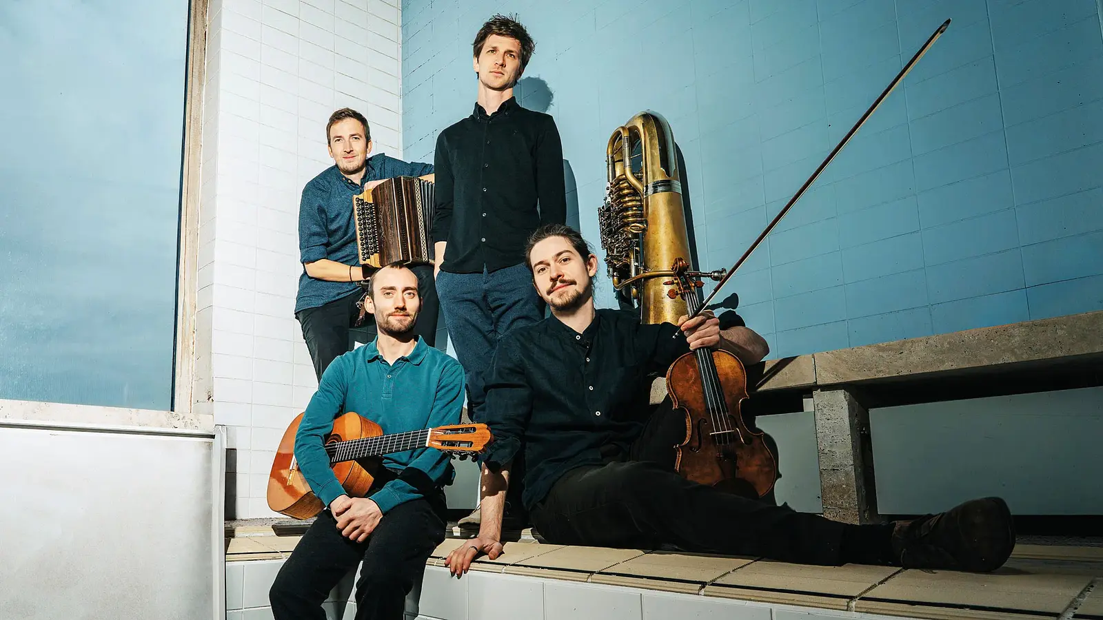 Vier Musiker mit Geige, Steirischer Harmonika, Gitarre und Tuba posieren in einem hellen Raum mit gefliesten Wänden