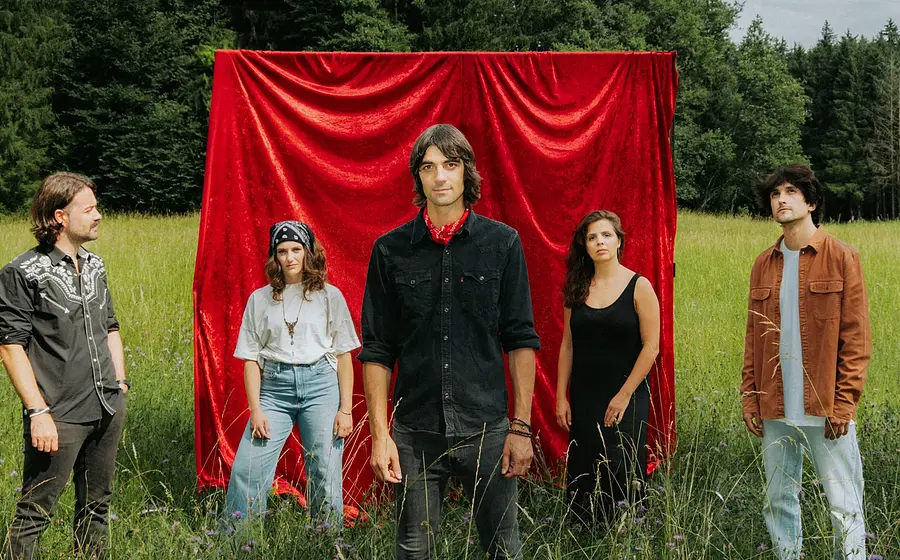 Foto für die Veranstaltung Max von Milland & Die Brennenden Herzen, mit der Band in der Natur und hinter ihnen einem roten Vorhang.