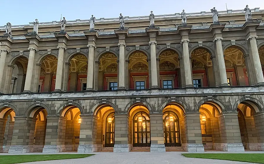 Ansicht eines historischen Gebäudes mit mehrgeschossiger Säulenfassade, Rundbogenarkaden und beleuchteten Eingängen im Erdgeschoss, aufgenommen vor hellem Abendhimmel.
