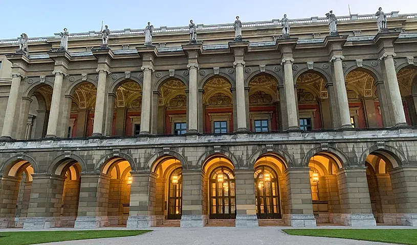 Ansicht eines historischen Gebäudes mit mehrgeschossiger Säulenfassade, Rundbogenarkaden und beleuchteten Eingängen im Erdgeschoss, aufgenommen vor hellem Abendhimmel.