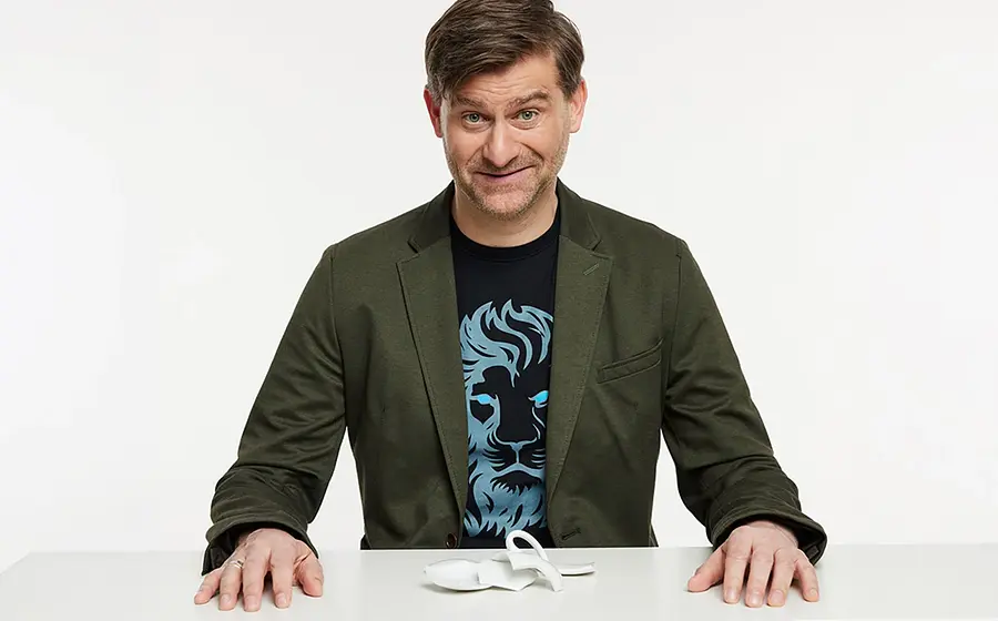 Michael Altinger an einem weißen Tisch vor hellem Hintergrund, in grünem Sakko und dunklem T Shirt mit Löwenmotiv, vor sich eine zerbrochene weiße Tasse auf der Tischplatte.