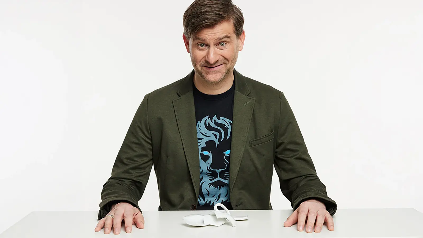 Michael Altinger an einem weißen Tisch vor hellem Hintergrund, in grünem Sakko und dunklem T Shirt mit Löwenmotiv, vor sich eine zerbrochene weiße Tasse auf der Tischplatte.