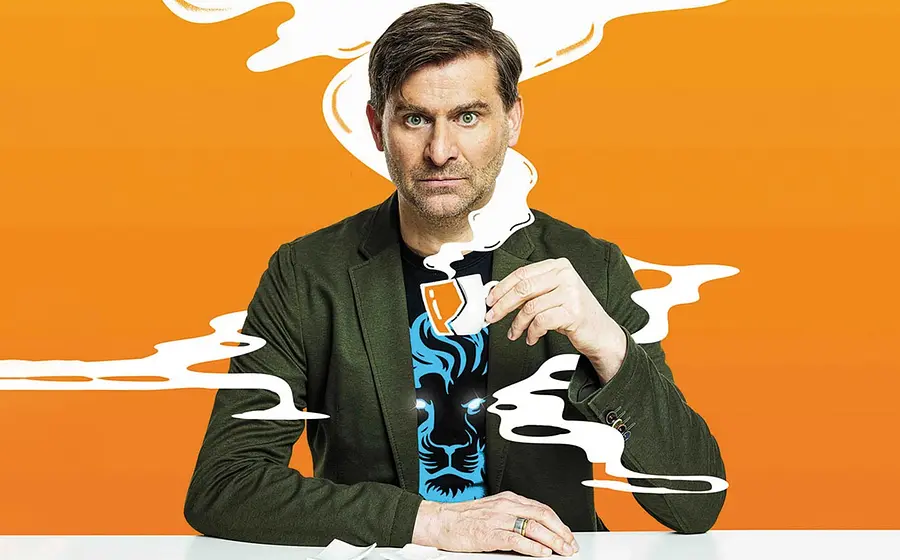 Porträtfoto von Michael Altinger in grünem Sakko und schwarzem T‑Shirt mit blauem Löwenmotiv vor orangefarbenem Hintergrund, sitzend an einem weißen Tisch und eine kleine Kaffeetasse in der Hand haltend, umgeben von grafisch dargestellten weißen Dampfschwaden.
