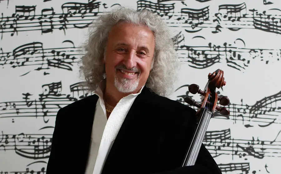 Promobild für das Event ‚Mischa Maisky, Violoncello‘: Eine Person mit lockigem, hellem Haar trägt dunkle Kleidung und hält ein Violoncello. Im Hintergrund sind großformatige, schwarz gedruckte Notenlinien und musikalische Zeichen zu sehen.