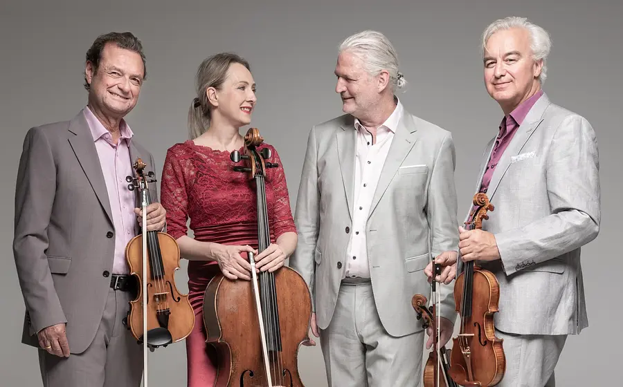 Das Modern String Quartetts mit vier Personen in eleganter Konzertkleidung vor hellgrauem Hintergrund, drei mit Violine und Violoncello in den Händen, eine Person im roten Spitzenkleid zwischen drei Personen in hellen Anzügen.