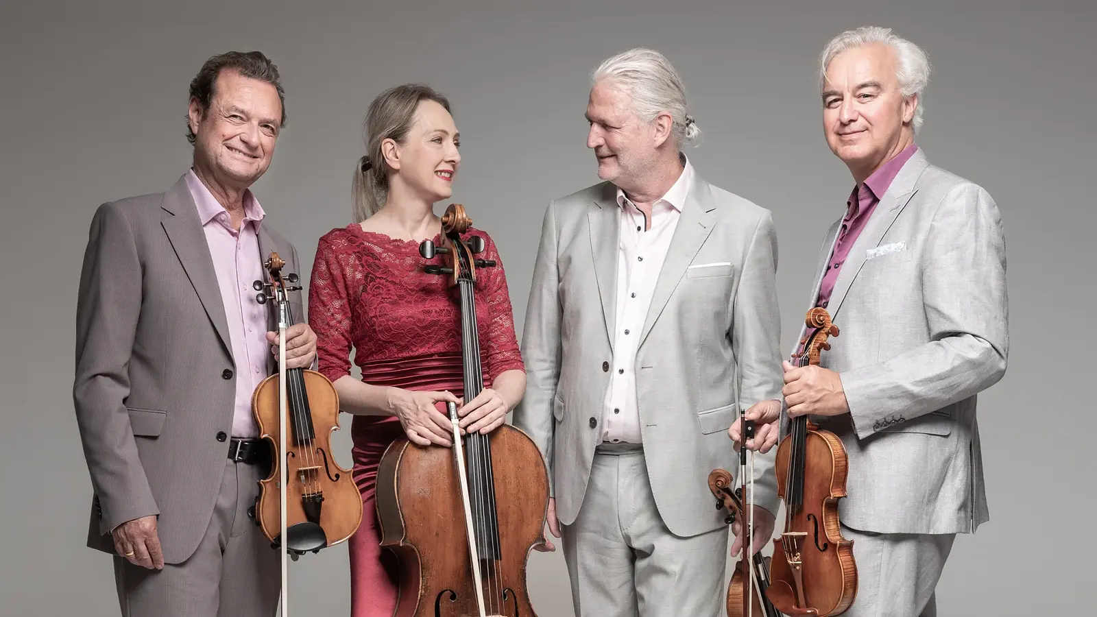 Das Modern String Quartetts mit vier Personen in eleganter Konzertkleidung vor hellgrauem Hintergrund, drei mit Violine und Violoncello in den Händen, eine Person im roten Spitzenkleid zwischen drei Personen in hellen Anzügen.