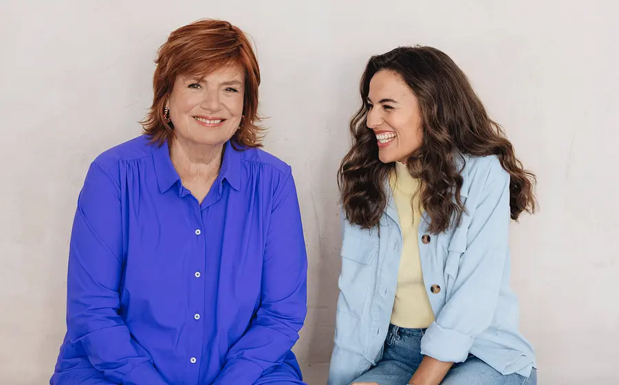 Portrait von Mona Ameziane und Christine Westermann vor hellem Hintergrund. Eine Person mit rotem Haar trägt eine blaue Bluse und blickt in die Kamera, während die andere Person in heller Jacke und Jeans seitlich zu der ersten Person gewandt ist.