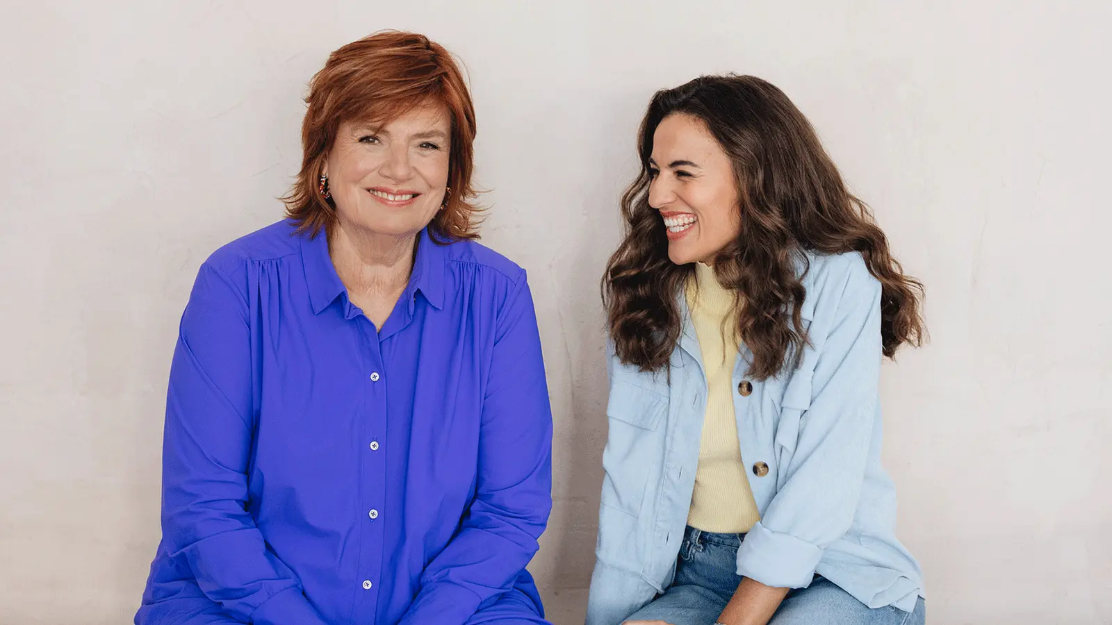 Portrait von Mona Ameziane und Christine Westermann vor hellem Hintergrund. Eine Person mit rotem Haar trägt eine blaue Bluse und blickt in die Kamera, während die andere Person in heller Jacke und Jeans seitlich zu der ersten Person gewandt ist.