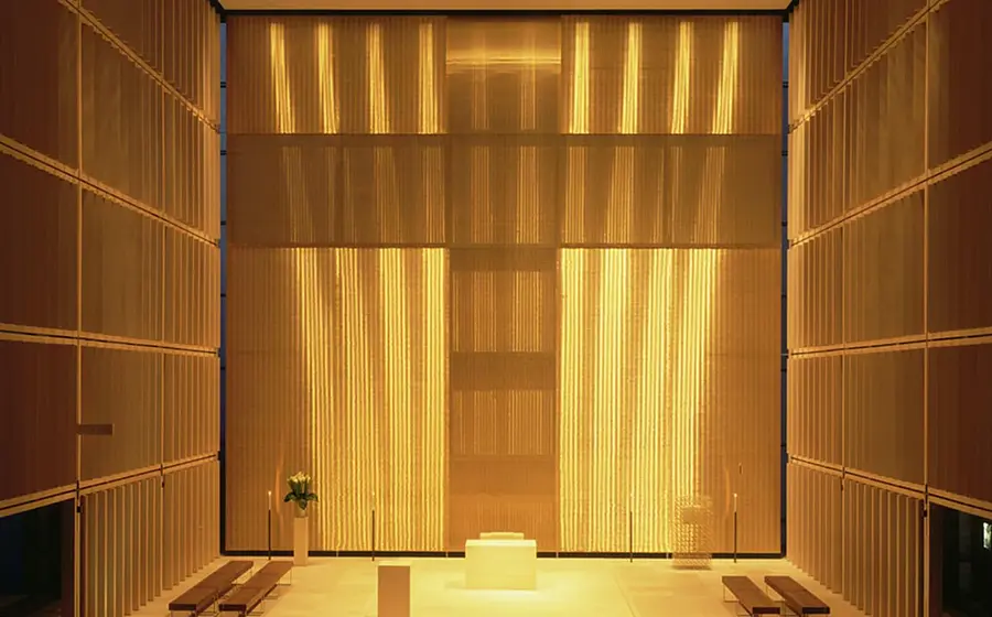 Innenansicht der Herz-Jesu-Kirche mit golden beleuchtetem Altarraum, zentralem Altarblock und hohen Lamellenwänden, die ein leuchtendes Kreuz im Hintergrund bilden.