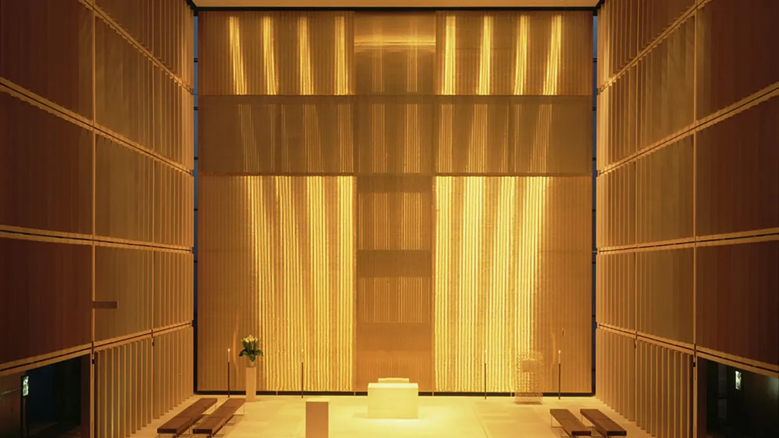 Innenansicht der Herz-Jesu-Kirche mit golden beleuchtetem Altarraum, zentralem Altarblock und hohen Lamellenwänden, die ein leuchtendes Kreuz im Hintergrund bilden.