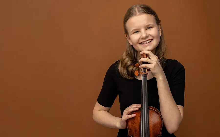 Eine jungen Person in schwarzem Oberteil vor warmbraunem Hintergrund, die eine Violine aufrecht hält und den Kopf auf den Wirbelkasten stützt, mit direktem Blick zur Kamera.