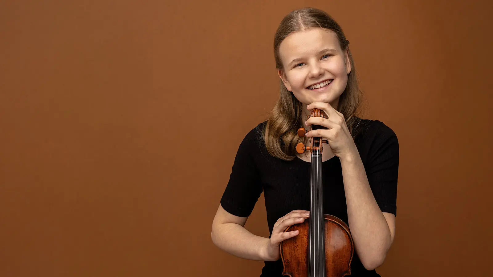 Eine jungen Person in schwarzem Oberteil vor warmbraunem Hintergrund, die eine Violine aufrecht hält und den Kopf auf den Wirbelkasten stützt, mit direktem Blick zur Kamera.
