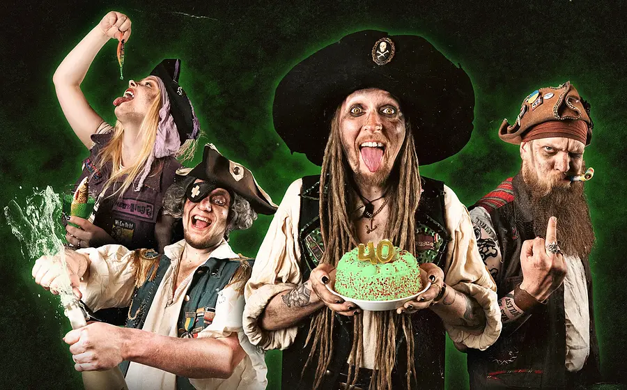 Vier Personen in Piratenkostümen vor grünem Hintergrund, eine Person hält eine Torte mit 40, andere mit Flasche und bunten Requisiten, Gruppenmotiv für Party‑Konzert