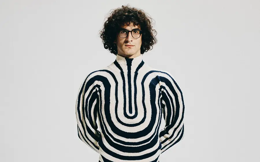 Giorgi Gigashvili mit lockigem dunklem Haar und runder Brille steht vor hellem grauem Hintergrund und trägt einen cremefarbenen Pullover mit schwarzen, symmetrischen Wellenmustern