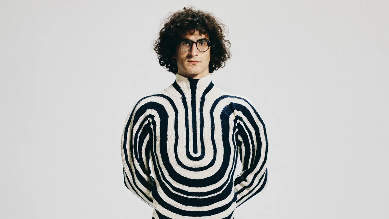 Giorgi Gigashvili mit lockigem dunklem Haar und runder Brille steht vor hellem grauem Hintergrund und trägt einen cremefarbenen Pullover mit schwarzen, symmetrischen Wellenmustern