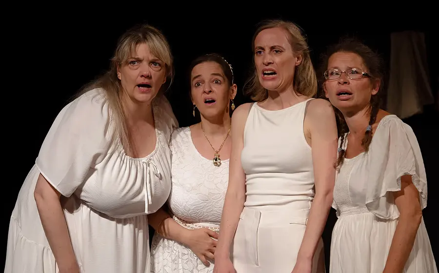 Bühnenfoto aus Lysistrata mit vier Darstellenden in weißen Kostümen vor schwarzem Bühnenhintergrund, die dicht beieinanderstehen und nach vorne blicken, eine Person mit Brille und drei weitere in unterschiedlichen weißen Kleidern